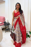 Woman wearing a red and white ikkat saree with a pattern, holding a handbag indoors.