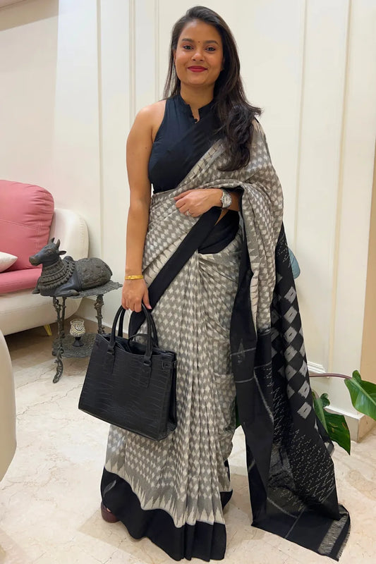 Woman in a black and grey ikkat saree holding a black handbag in an indoor setting