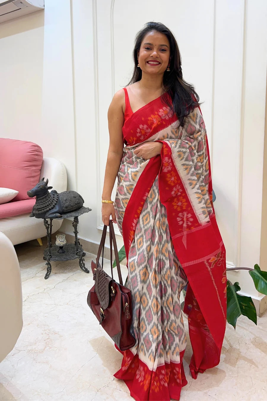 Woman wearing a red and white ikkat saree with a pattern, holding a handbag indoors.