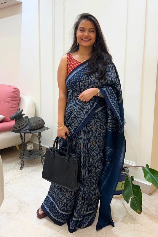 A woman in a blue handloom ikkat saree with a red blouse, holding a black handbag indoors.