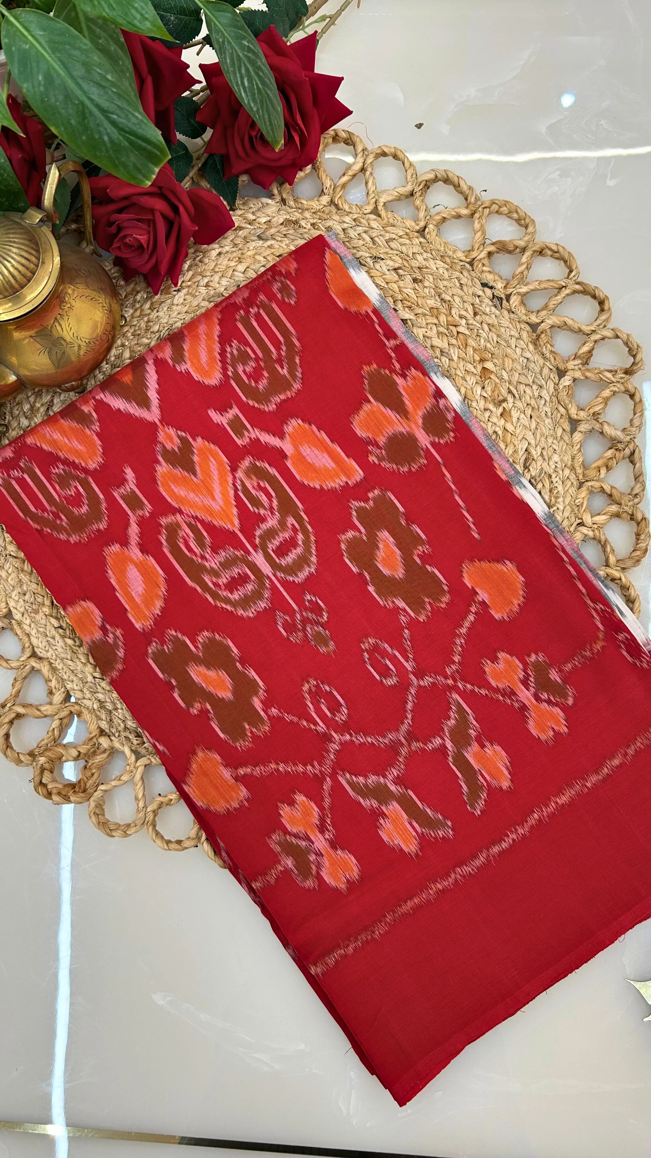 Red ikkat saree with floral patterns on a decorative mat with roses and a lamp.
